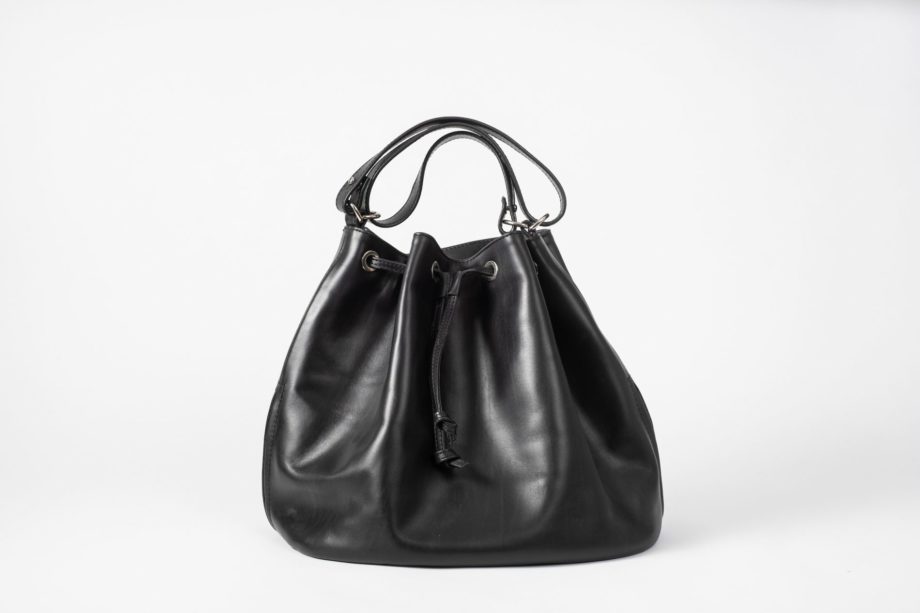 Front view on a black vegetable tanned leather Bucket Bag with a short handle folded three times and a string passing the eyelets and hanging in the front of the bag